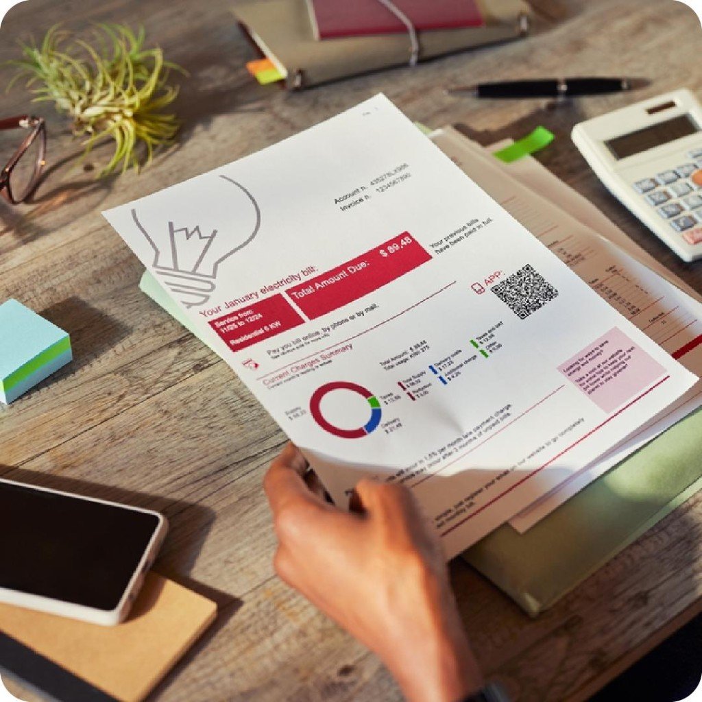 A hand holding a residential electricity bill above a desk with a calculator and phone.