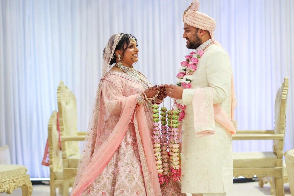 A bride and groom in pink and cream wedding clothes exchanging flower garlands at a wedding ceremony.