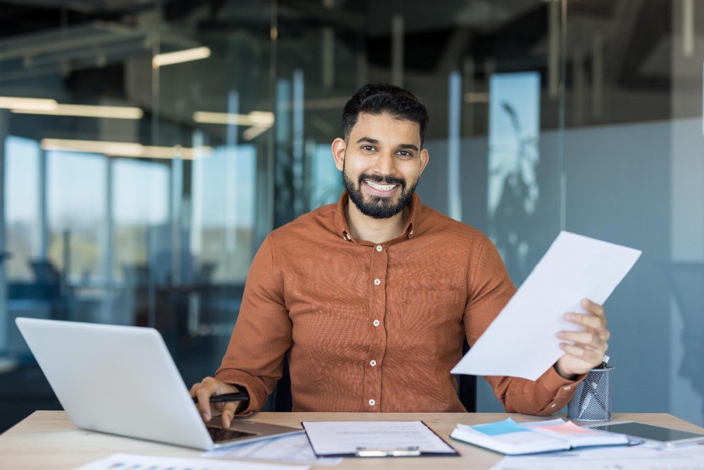 A business owner holding registration papers at a desk with a laptop.